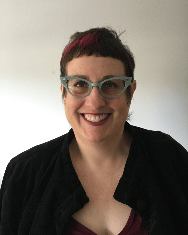 Portrait of a smiling woman with short dark hair and a pink streak, wearing teal cat-eye glasses and a black top, photographed against a plain background.